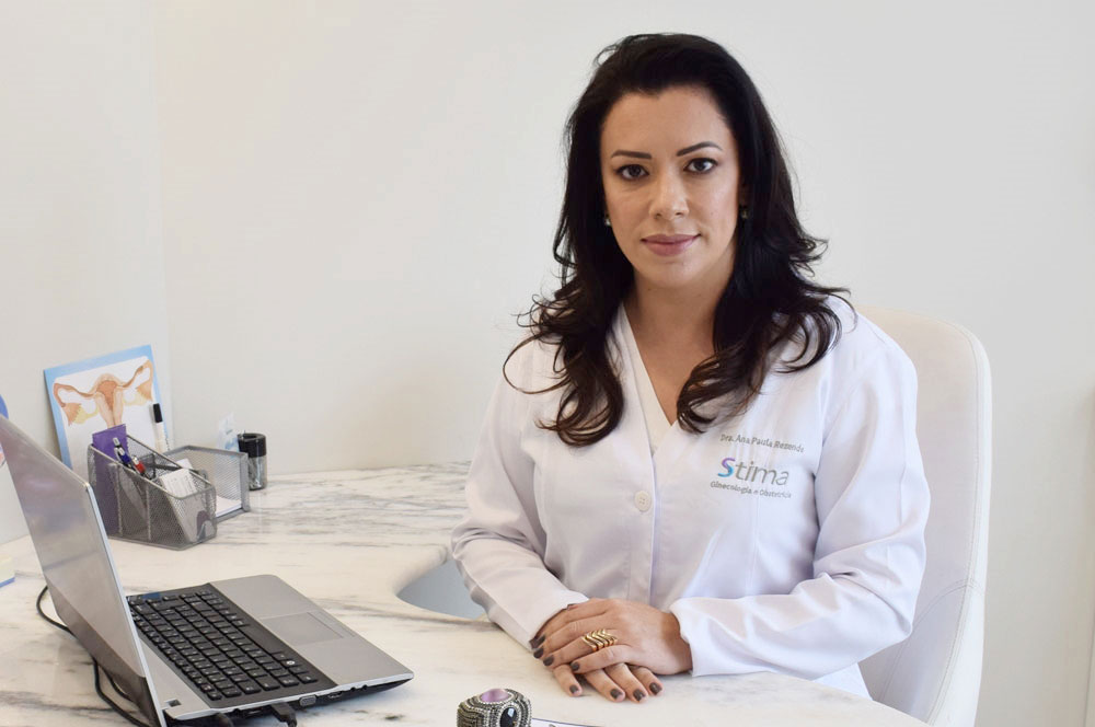 Dra Ana Paula Rezende: Mulher, cabelos pretos longos, vestindo jaleco, mãos apoiadas sobre a mesa. Olhando para a câmera.